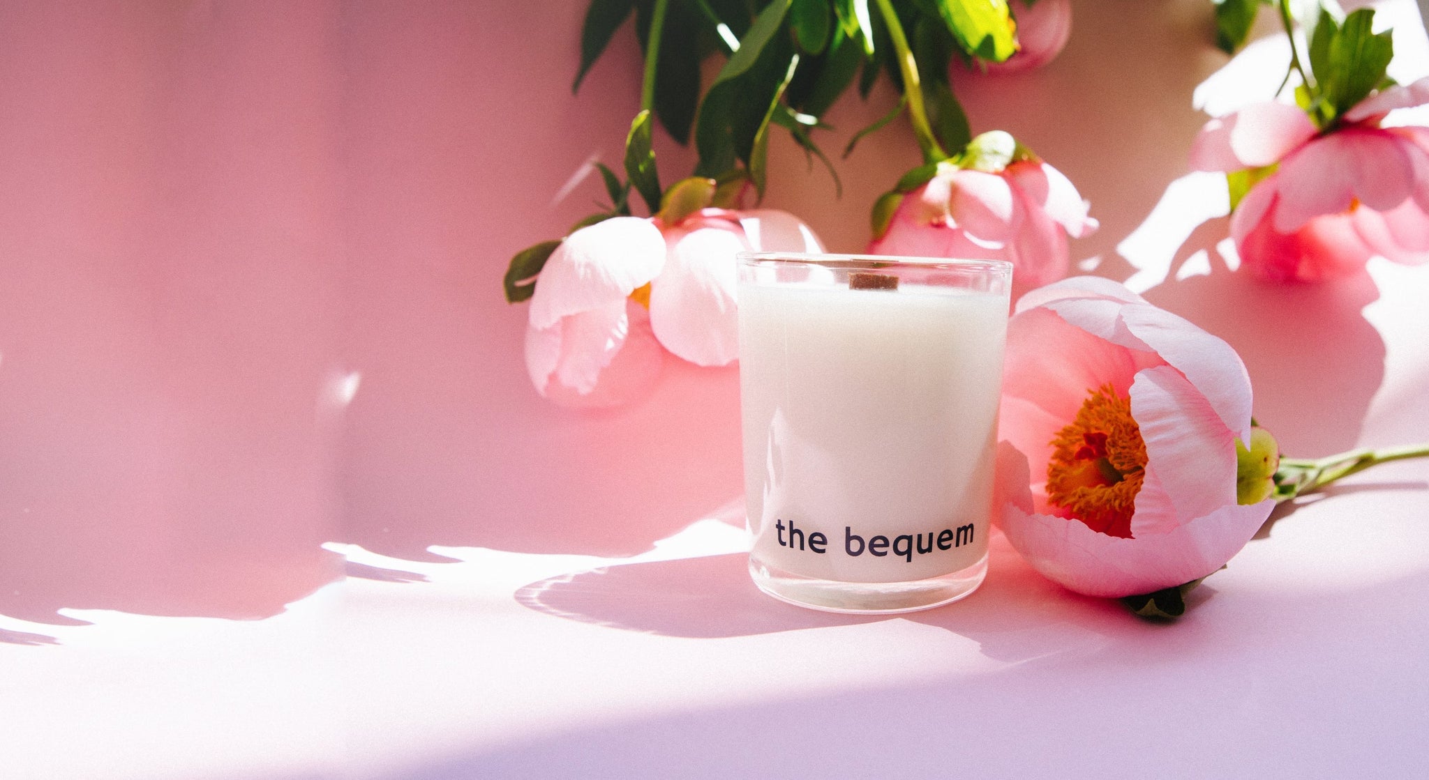 The Bequem candle placed among pink peonies on a sunlit surface, creating a soft and romantic atmosphere that evokes springtime freshness.