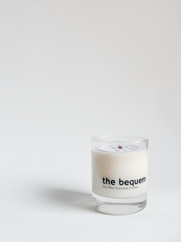 A white candle with 'the bequem' printed on it, placed in a clear glass holder against a light background.