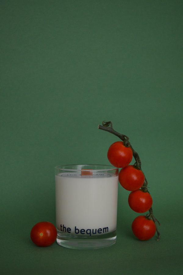 A summer garden soy wax candle with a wooden wick, in a clear glass container, labelled 'The Bequem', surrounded by cherry tomatoes on a green background.