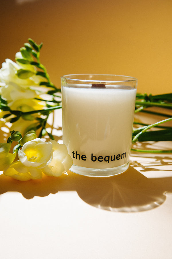 The Bequem white soy wax candle with a wooden wick, placed on a yellow surface and surrounded by white freesias and green leaves under warm natural sunlight