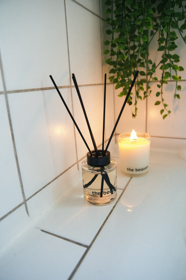 The Bequem reed diffuser and soy candle set displayed on white tiles with green foliage and soft candlelight for a cozy home spa atmosphere