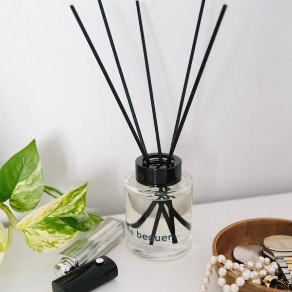 Reed diffuser by The Bequem placed on white surface beside green plant, perfume bottle, and jewelry for lifestyle styling.