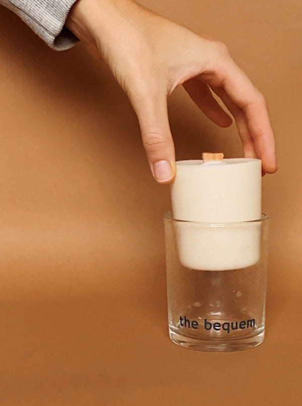 A hand placing a white candle refill into a clear glass candle holder with the brand name 'the bequem' visible on the glass.