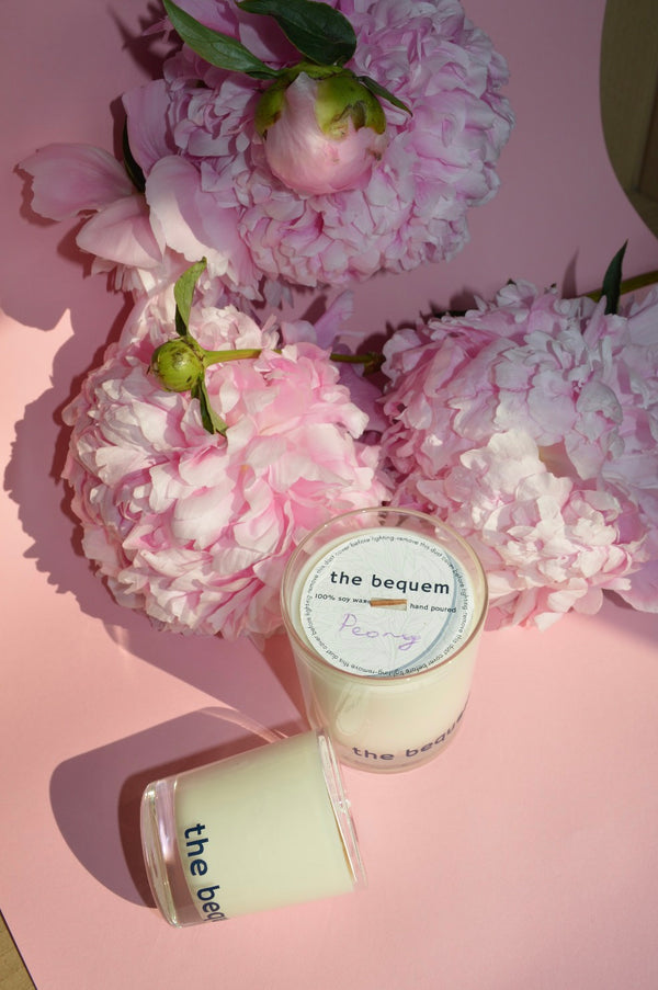 The Bequem Peony soy wax candle in a clear glass jar with a wooden wick, styled on a soft pink surface surrounded by blooming peony flowers for a fresh floral aesthetic